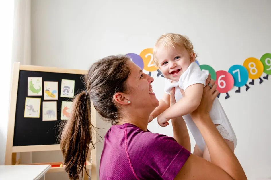 Professora somrient aixecant un bebe en una aula d'una escola infantil
